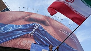 رجل يلوّح بعلم إيران خلال حملة مؤيدة للحكومة تحت لافتة في وسط طهران تُظهر مضيق هرمز وشفاه الرئيس الأمريكي دونالد ترامب مخيطة