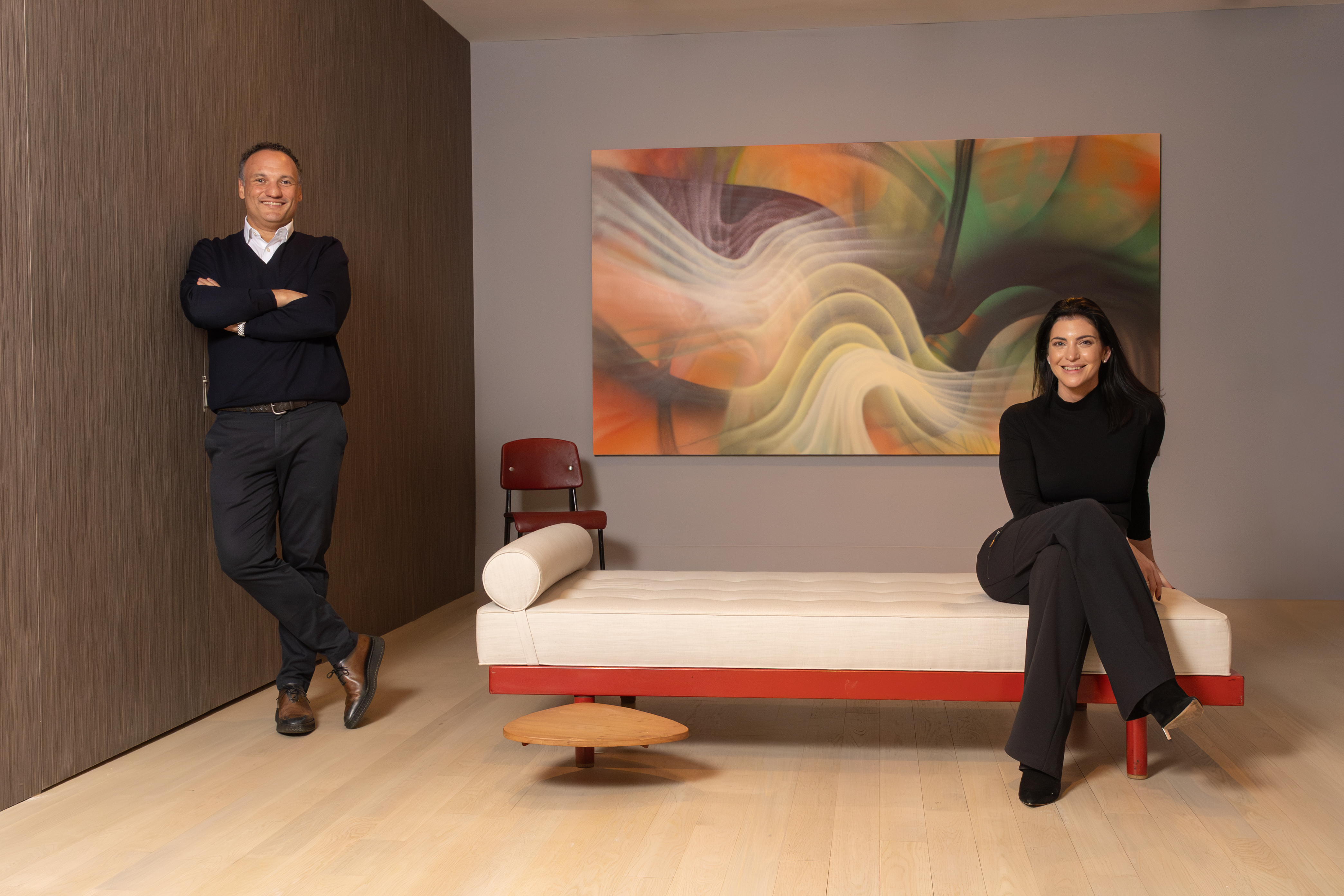 Two people pose in a modern room with a long light-colored daybed, standing and sitting beneath a large abstract painting of soft, wavelike shapes.