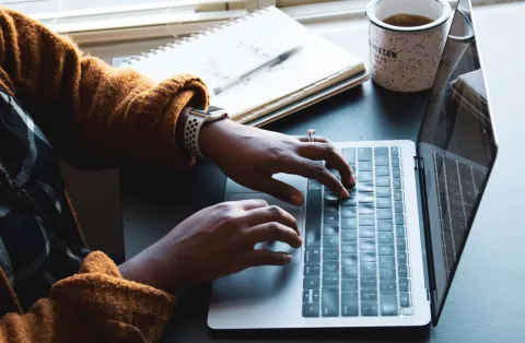 Fingers typing on a laptop with notepad, pen and mug in the background