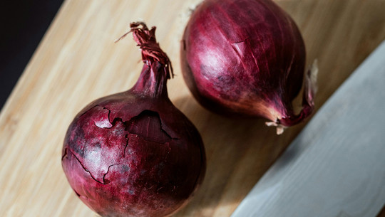 Warum wir alle öfter rote Zwiebeln essen sollten Zwei rote Zwiebeln mit teilweise abblätternder Schale liegen auf einem hellen Holzbrett.