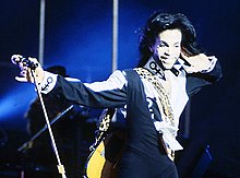 Waist high portrait of thirty year old male, wearing longish hair and short beard, and a black jumpsuit over one shoulder, right hand extended to hold microphone stand. Shirt has unnamed "Prince" symbol on it. Guitar visible behind him.