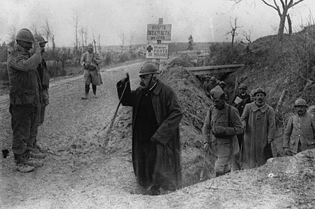 Photo en noir et blanc d'une personne aux cheveux blancs, à la moustache foncée et avec un casque d'artilleur sortant d'une tranchée et saluant deux militaires