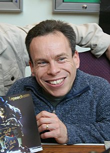 Actor Warwick Davis sits at a table, smiling and holding Leprechaun merchandise.