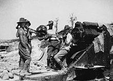 Bare-chested men in slouch hats and breeches stand by a large artillery piece, ready to load a shell.