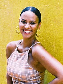 Against a yellow wall outdoors, a woman with hoop earrings and a plaid summer top smiles widely, her dark hair pressed tightly against her scalp.