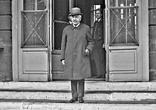 Photo en noir et blanc d'un homme se tenant sur les marches d'un bâtiment, avec chapeau melon, sourcils noirs, lorgnons, moustache grise, manteau gris