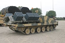 vehicle facing right in desert camouflage 120 mine laying dispensers on the flatbed part of the vehicle. To the rear is another armoured vehicle carrying two metal Fascine