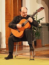 Photo couleur d'un homme tout de noir vêtu, assis, de face, en train de jouer de la guitare.