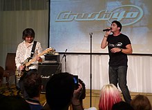 A Japanese man (left) plays a guitar while an American man (right) sings in front of a crowd.