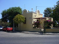 Durrell's house in Rhodes features Mediterranean architecture and has yellow-painted stucco or plaster walls. It is located on a paved asphalt street, with two cars parked parallel to it. The house is surrounded by several trees, shrubbery, roses, and flowering bushes.