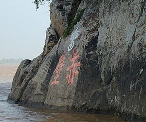 A photograph of some cliffs above a waterline, the cliffs have two red Chinese characters painted at their centre with a white Chinese character at the top centre of the red ones