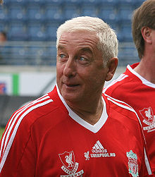 The head and upper torso of a gentleman in his late 50s. He has short white hair and is wearing a red football shirt, which has the Liverpool FC crest on the left breast, and a crest on the right breast that says "Liverpool Legends". A white logo of the Adidas sponsor is visible in the centre of the shirt, and three white stripes are present on the shoulder.