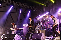 A rock band performing live on-stage. The stage is brightly lit on the left, a man plays bass behind a keyboard. In the centre, a man plays guitar and behind him a man plays drums. On the right, a man plays an electric mandolin.