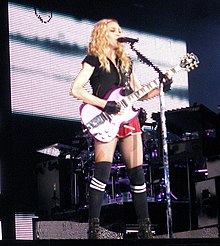 Picture of a blond woman performing on a stage. She appears to wear red shorts and plays as pink guitar and sings to a microphone. Behind her, there are video screens showing horizontal graphical patterns.