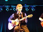 Rodney Crowell, playing an acoustic guitar while seated on a stool.