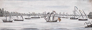 A group of 14 small ships of varying sizes on a lake with wooded land in the background