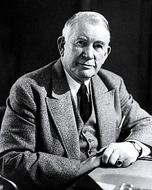 A man with white hair wearing a gray jacket and vest, black tie, and white shirt, seated and leaning on a desk