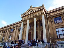 The facade of a masonry building, with four Greek adorning its entrance, under a clear blue sky