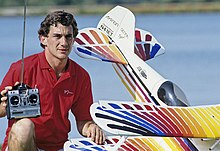 Photo d'Ayrton Senna en polor rouge qui prend la pose avec un avion miniature téléguidé, télécommande à la main.