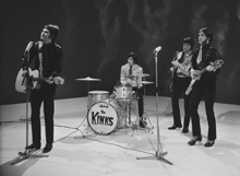 Four members of the band the Kinks stand onstage, during a television appearance. From left to right is Ray Davies, wearing a leather suit and long black pants. He is strumming an acoustic guitar and preparing to sing into a microphone placed in front of him. Next is Mick Avory, seated and playing the drums. He is wearing a brightly coloured long-sleeved shirt and dark pants. After him is bassist Pete Quaife, playing his instrument and wearing a set of clothes similar to Avory's. Last is Dave Davies, wearing what appears to be a leather suit with a short tie, black pants and high-heeled loafers. He plays an elaborate v-shaped electric guitar and is standing behind a microphone.