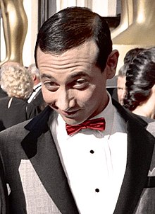 Photographie du buste d'un homme au cheveux gominés et au sourire mystérieux. Il porte un smoking gris et un nœud papillon rouge.