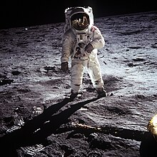 An astronaut in an American Apollo-program spacesuit, standing on the flat, heavily footprinted landing area, with the utterly black sky of space above the horizon.