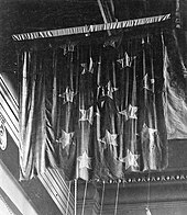 A black-and-white photo of a battle-worn flag suspended from the ceiling of a museum