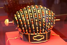 Photo couleur d'une couronne noire en forme de toque, ornée de perles de couleurs rouge, bleue et dorée. L'objet est posé sur un présentoir rouge dans un musée.