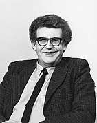 A curly-haired middle-aged man with a woolen suit, a narrow tie, and thick horn-rimmed glasses