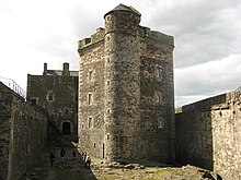 View of castle within its courtyard with a stone building over three stories tall in the middle. The building is flanked by stone walls on both sides that are half as tall.