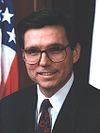 Hispanic man with large glasses and black hair with the US flag behind him