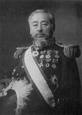 Portrait noir et blanc d'un homme âgé, en uniforme d'allure militaire sur fond sombre et portant une écharpe claire en bandoulière et, sur sa poitrine, des médailles.