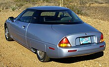 The rear three-quarters view of an electric automobile with a hidden rear wheel and a shown front wheel. Photo is taken in what looks like a desert