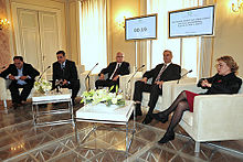 Four men and a woman sitting in white armchairs with microphones