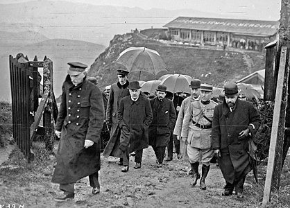 Photo en noir et blanc de plusieurs hommes (civils et militaires) arrivant au sommet d'une colline par un temps pluvieux ; au centre du cliché, un homme aux cheveux blancs, aux sourcils épais et foncés, à la moustache grise et portant un manteau de couleur sombre regarde l’objectif