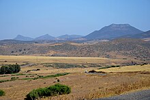 Rolling hills with mountains in the background, a road and electricity pylons cross the image from left to right. The ground is stony with some browned grass, a few bushes and scattered trees. A small farm is in the right mid-ground with some fields used for hay and grazing of sheep.