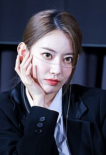 Sakura Miyawaki poses with her hand on her face, wearing a black blazer and decorative chain across her face.