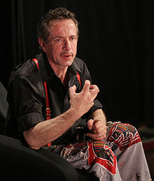 La photo montre Clive Barker parlant avec animation pendant une conférence. Il porte une chemise et un pantalon sombres et des bretelles rouges.