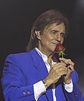 A imagem mostra o cantor Roberto Carlos, um homem de mais de 70 anos e branco, segurando uma rosa com as mãos, como ele costuma fazer antes de jogá-las para o público em seus shows. Ele está com um paletó azul.