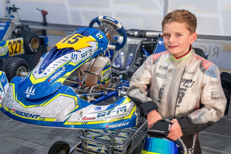 Ten-year-old Daniel Ferguson in his racing suit, holding a helmet, stands next to a blue and yellow go-kart.