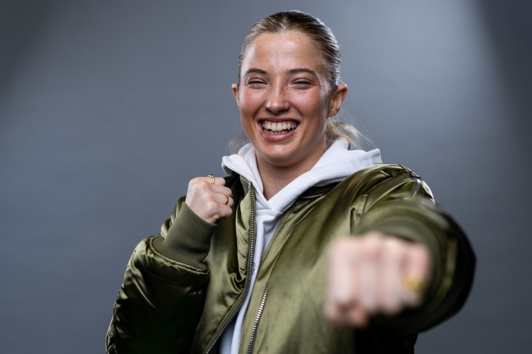 Fran Hennessy, a female boxer, smiling with closed eyes, wearing a white hoodie and a green bomber jacket, with one hand clenched and the other outstretched as if punching.