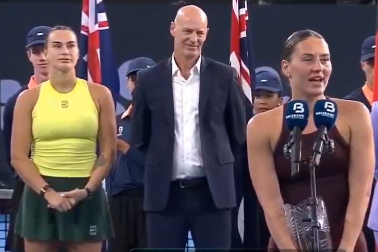 Marta Kostyuk, dressed in a yellow top and green skirt, stands next to Aryna Sabalenka, who is speaking at a podium with two microphones, after the Brisbane final.