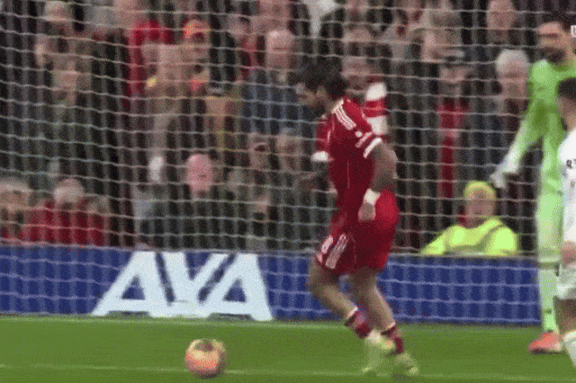 A soccer player in a red jersey attempts to kick a ball into the goal, but trips and falls.