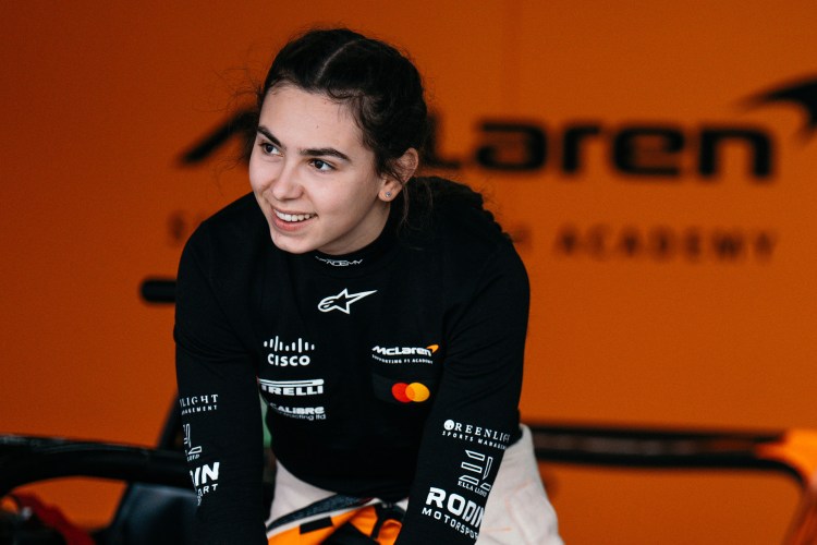 Ella Lloyd of Rodin Motorsport in a black long-sleeved shirt with various sponsor logos, including McLaren, Cisco, and Pirelli, looking on in the garage.