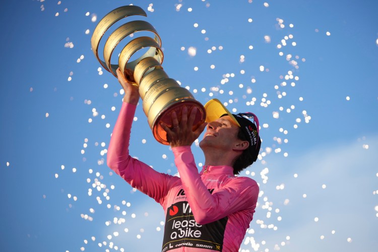 Adam Yates and Simon Yates smiling as they ride their bicycles, Simon wearing a pink jersey and Adam wearing a white jersey.